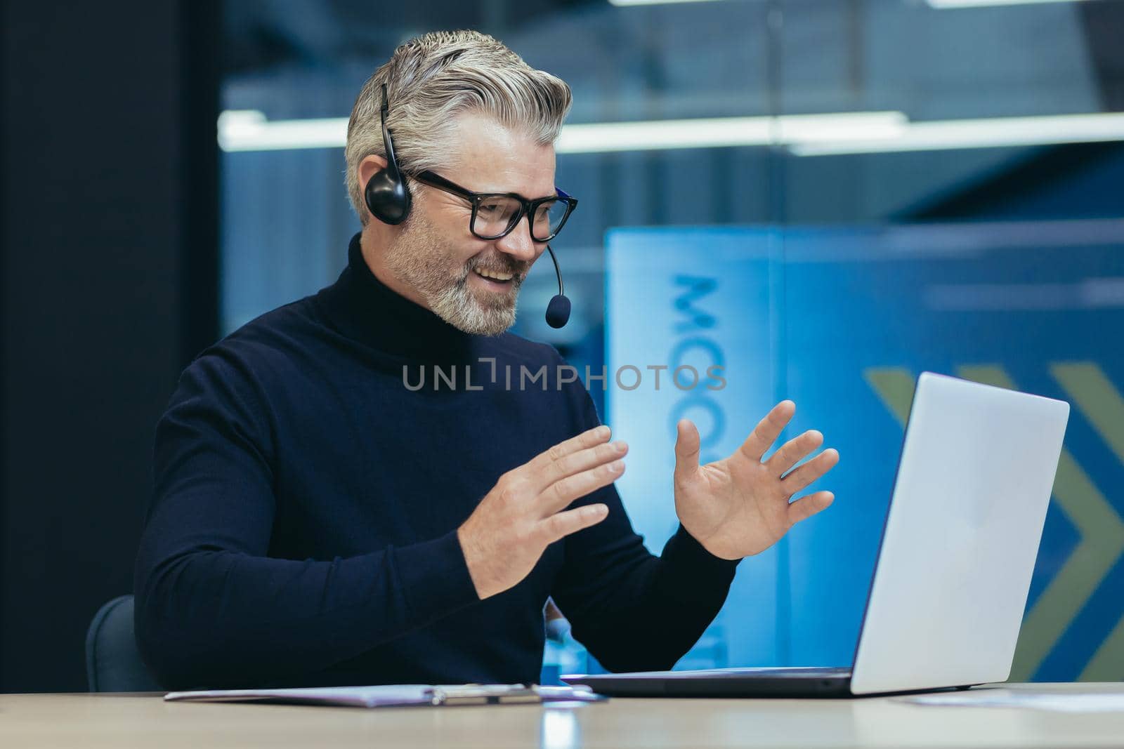 Senior experienced gray-haired businessman with headset, using laptop for online meeting, video call with colleagues businessman mature working inside modern office building.