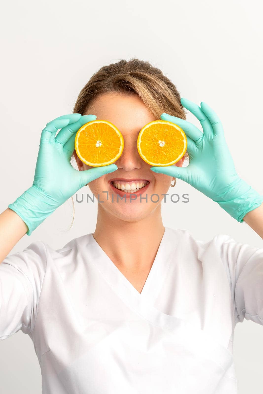 Portrait of young caucasian smiling female beautician covering eye with two orange slices wearing gloves over a white background