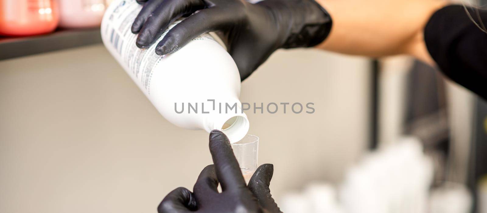 A hairdresser in black gloves is preparing hair dye with a bottle in a hair salon, close up. by okskukuruza