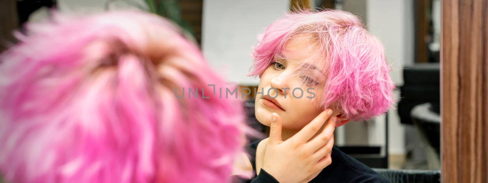 The beautiful young caucasian woman with a new short pink hairstyle looking at her reflection in the mirror checking hairstyle in a hairdresser salon. by okskukuruza
