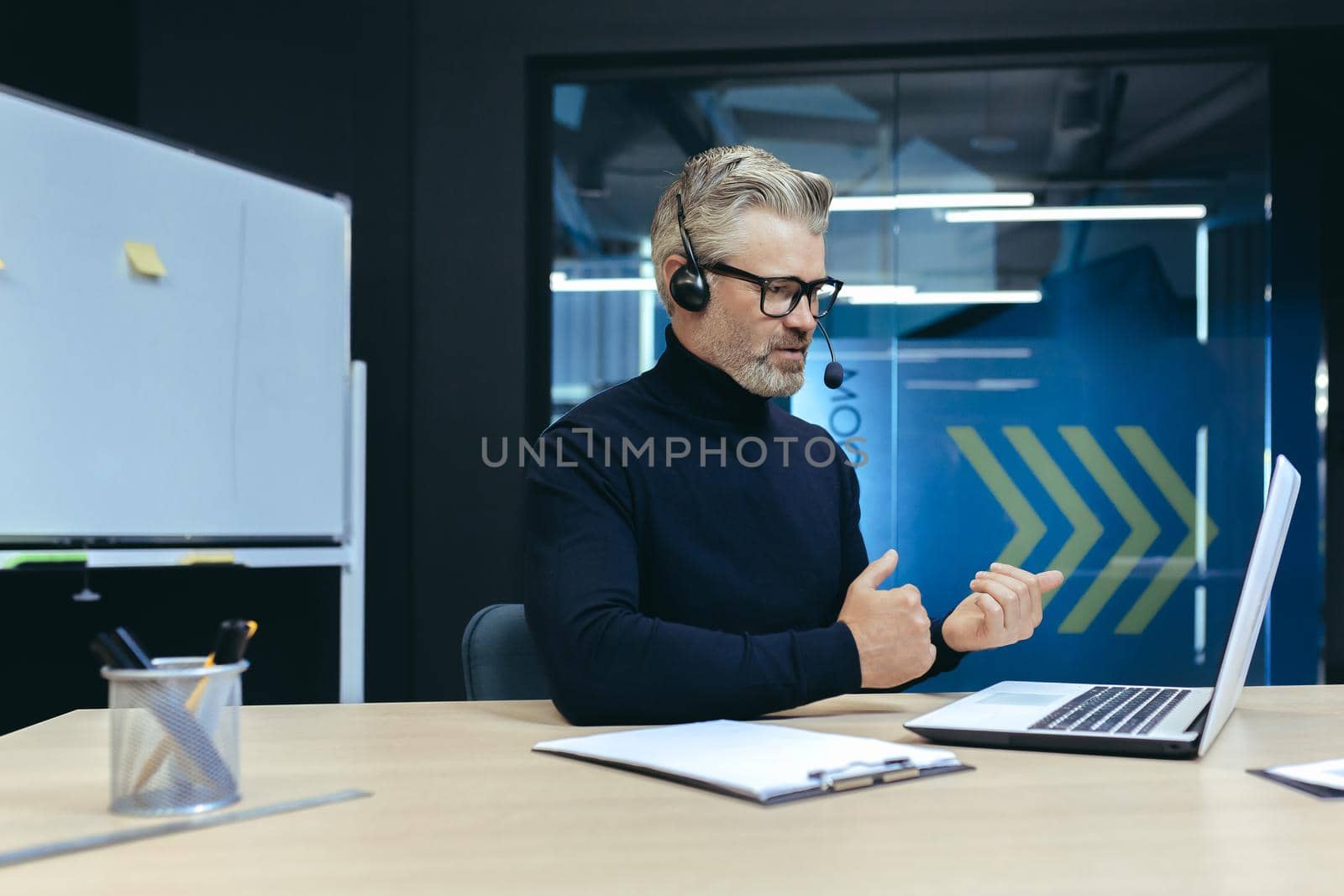Senior experienced gray-haired businessman with headset, using laptop for online meeting, video call with colleagues businessman mature working inside modern office building by voronaman