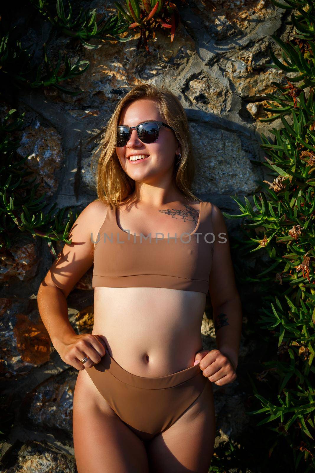 Beautiful girl in sunglasses in a beige swimsuit. Young woman with tanned body soit near stone wall with green plants