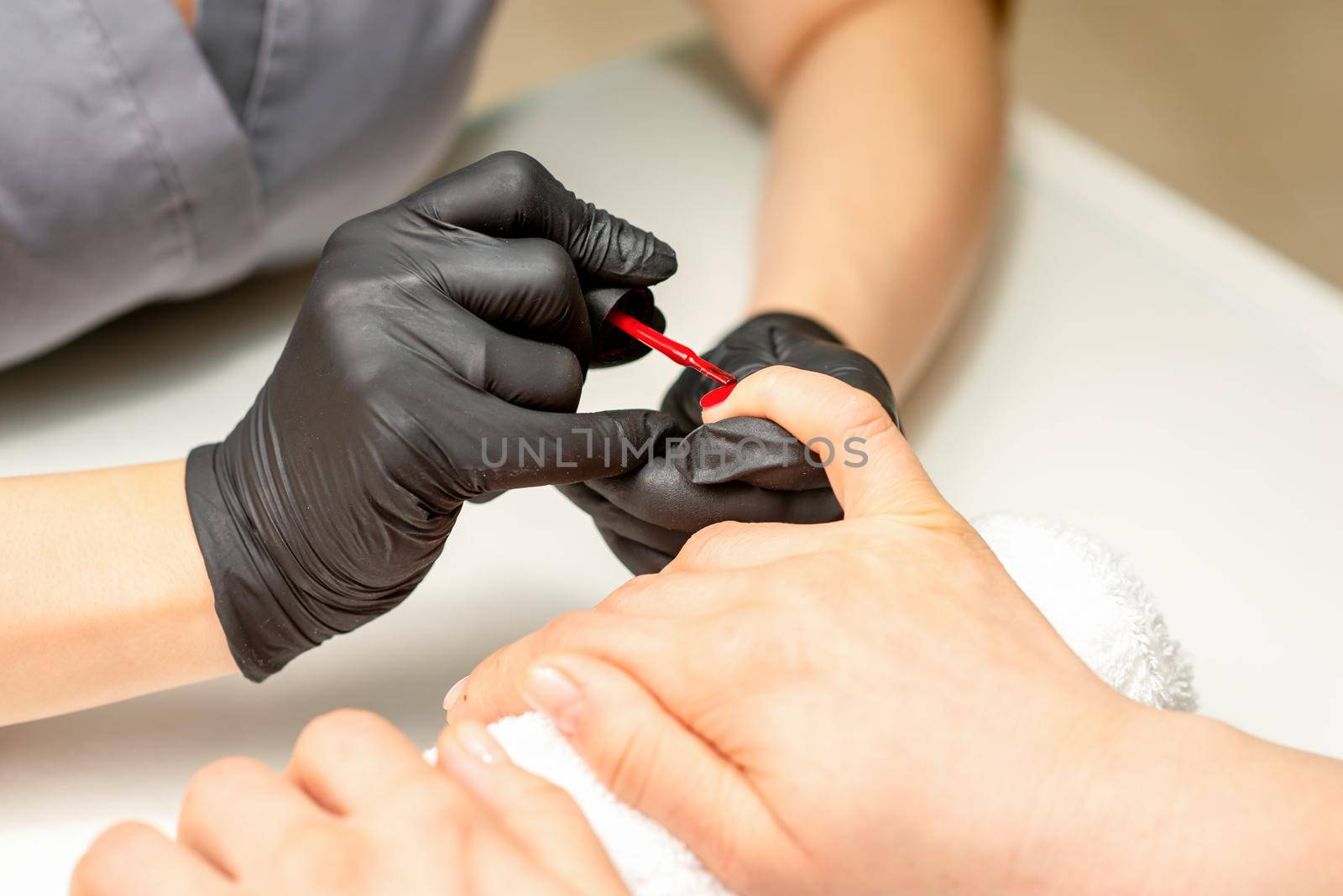 Manicure varnish painting. Close-up of a manicure master wearing rubber black gloves applying red varnish on a female fingernail in the beauty salon. by okskukuruza