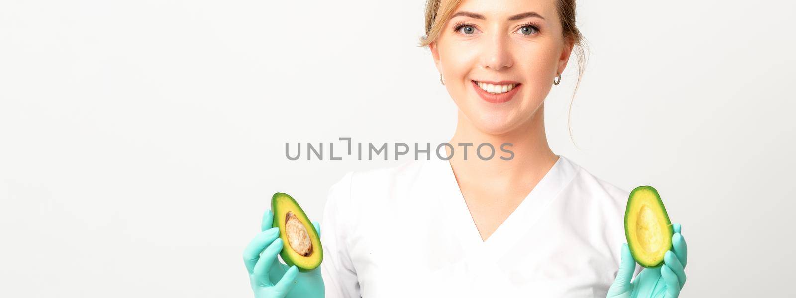 Portrait of smiling young female nutritionist doctor with organic avocado fruits posing at camera on white background, copy space. Benefits of proper nutrition