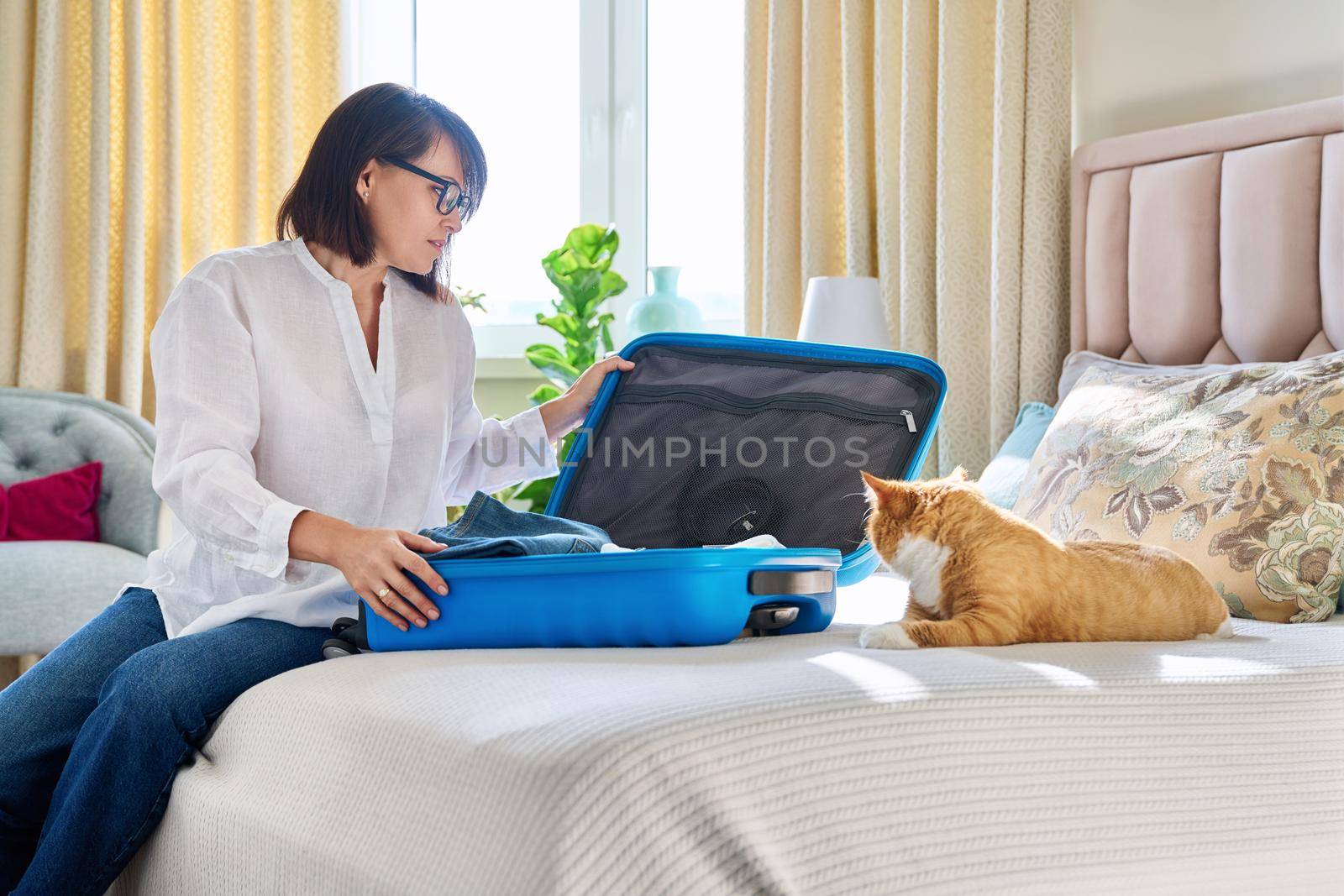 Happy woman packing luggage into suitcase with pet cat at home in bedroom. Mature female owner of old red pet going on trip, travel, vacation. Journey adventure holidays tourism concept