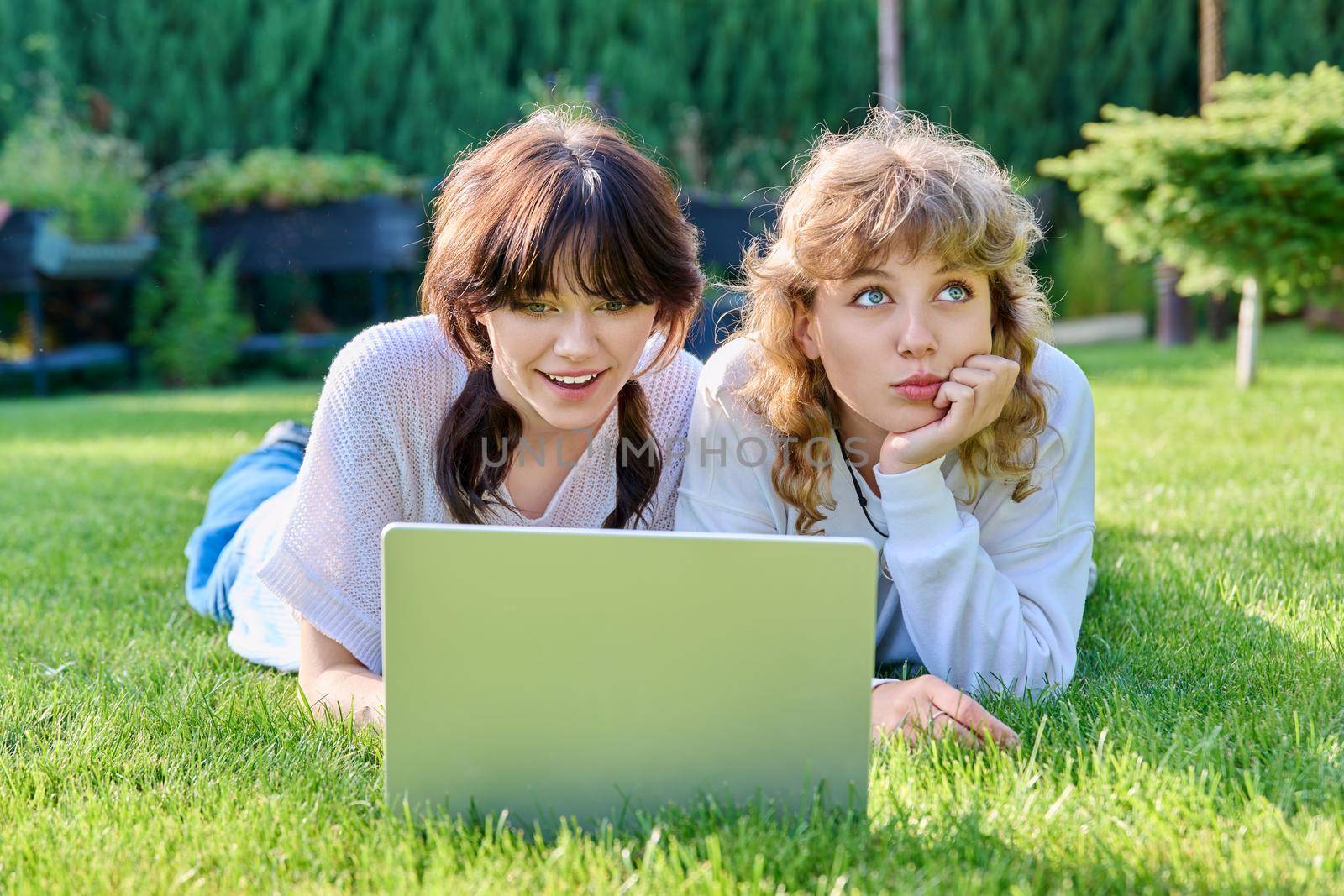 Two teenage girlfriends using laptop lying on grass on sunny summer day by VH-studio