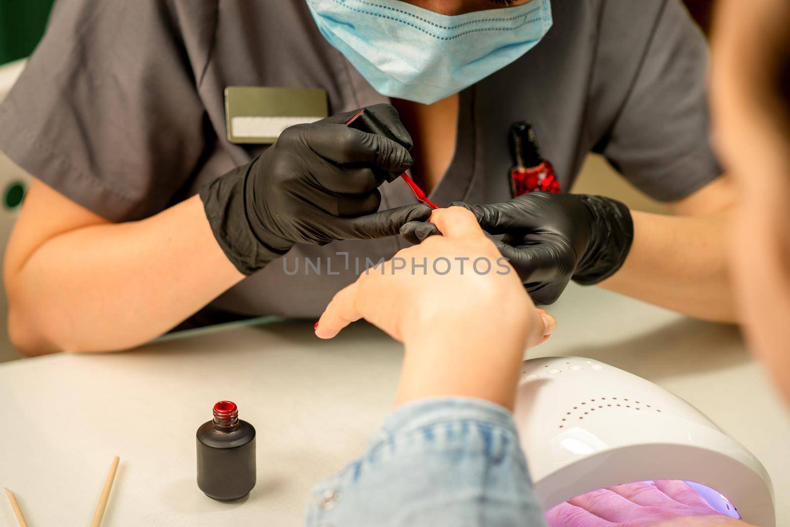 Manicure varnish painting. Close-up of a manicure master wearing rubber black gloves and a protective mask applying red varnish on a female fingernail in the beauty salon. by okskukuruza