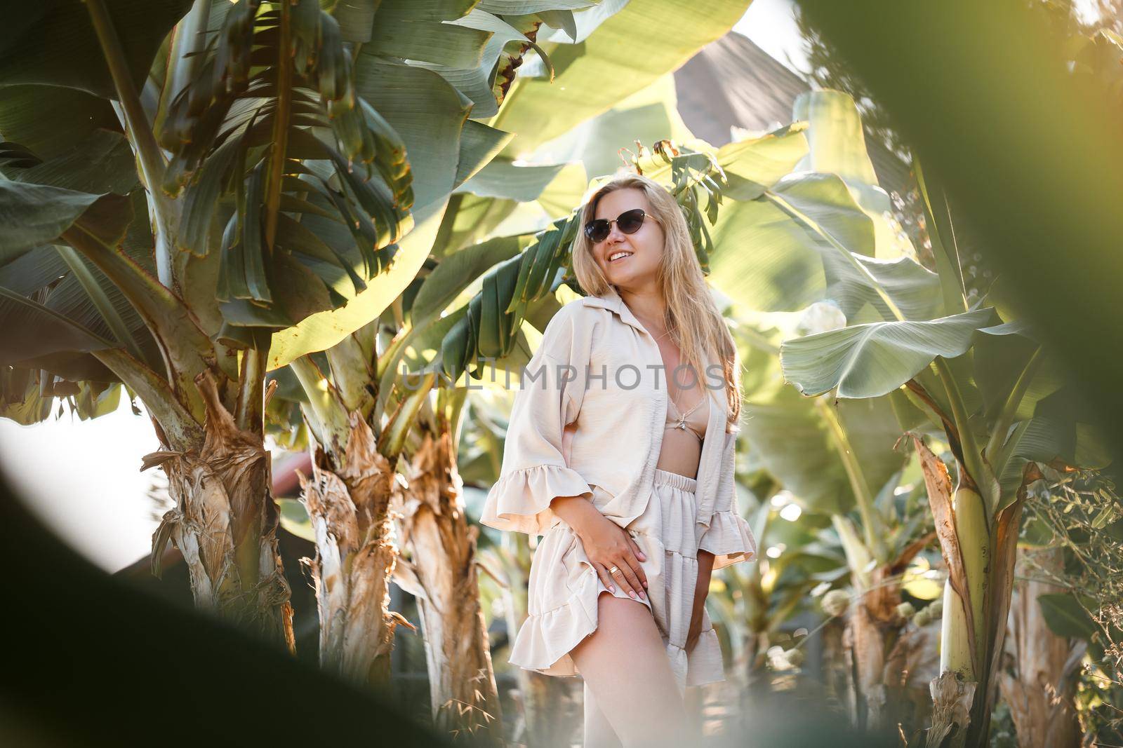 A beautiful young woman with long blond hair of European appearance stands near the banana trees. Girl in tropical forest on a sunny summer day. Selective focus by Dmitrytph