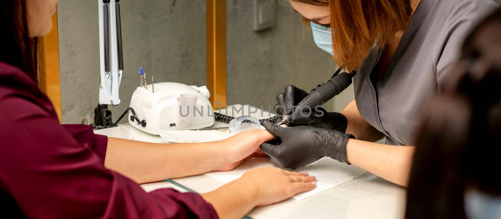 Manicure master wearing protective mask uses electric nail file machine in a nail salon, close up