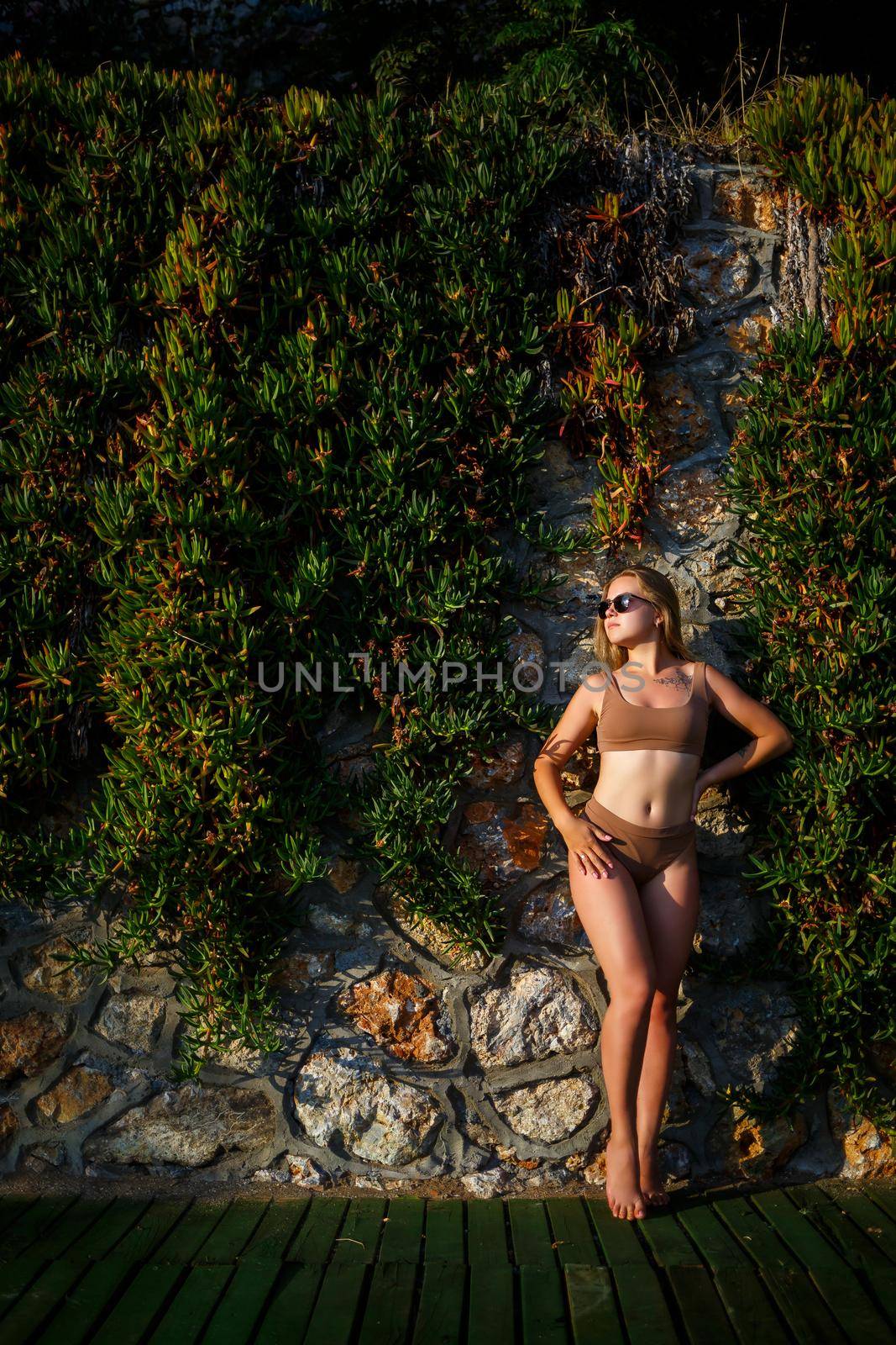 Beautiful sexy girl in sunglasses in a beige swimsuit. Young woman with tanned body soit near stone wall with green plants