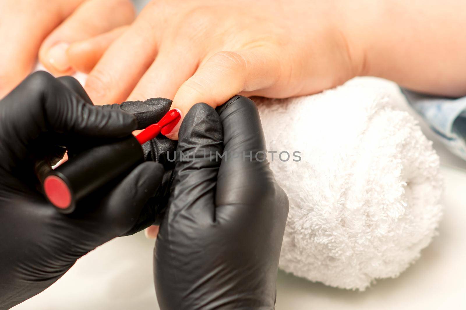 Manicure varnish painting. Close-up of a manicure master wearing rubber black gloves applying red varnish on a female fingernail in the beauty salon