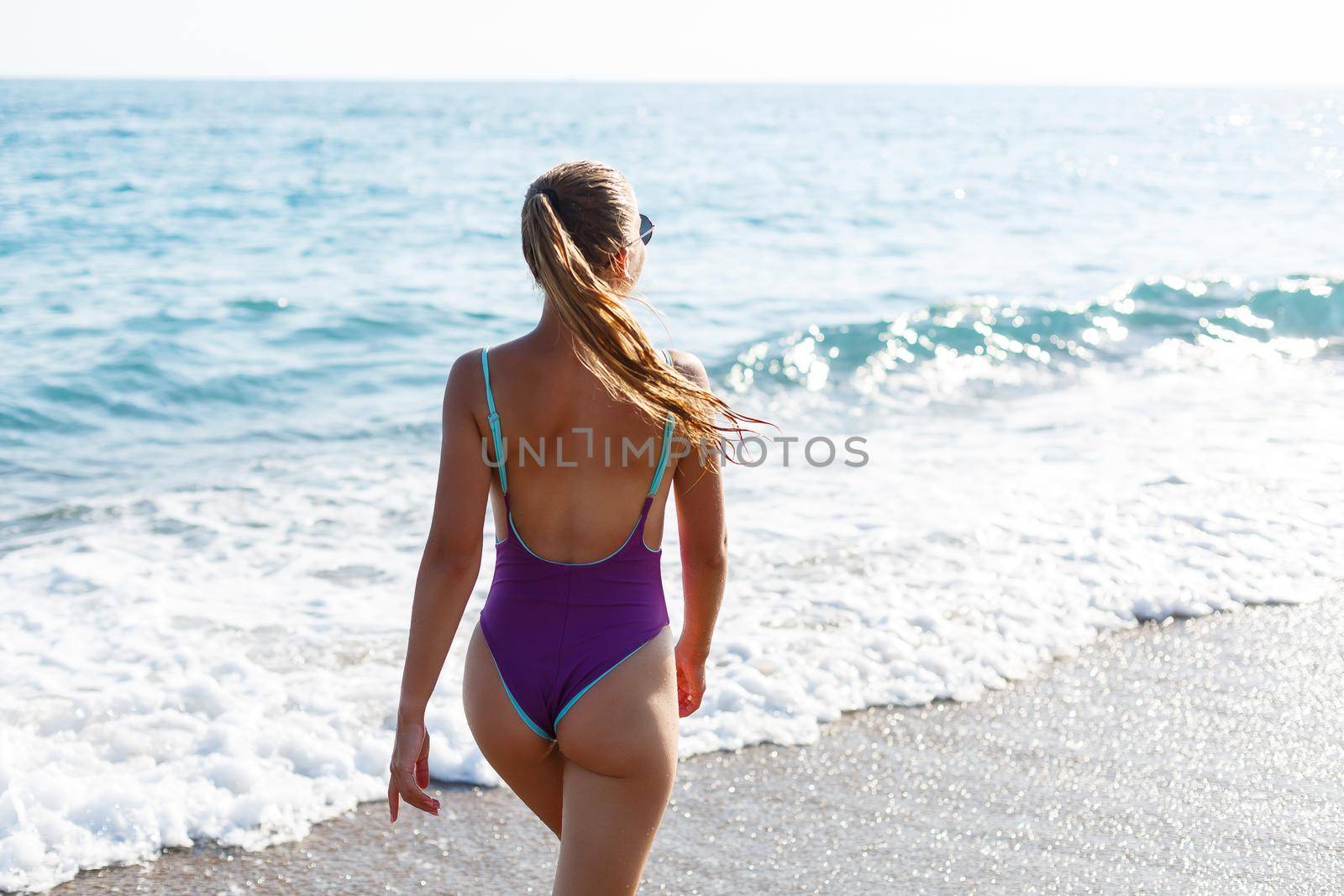 An attractive young woman comes out of the sea in a swimsuit. A girl in a swimsuit walks under the bright summer sun along the sand of the Mediterranean coast. Rest by the sea. Selective focus