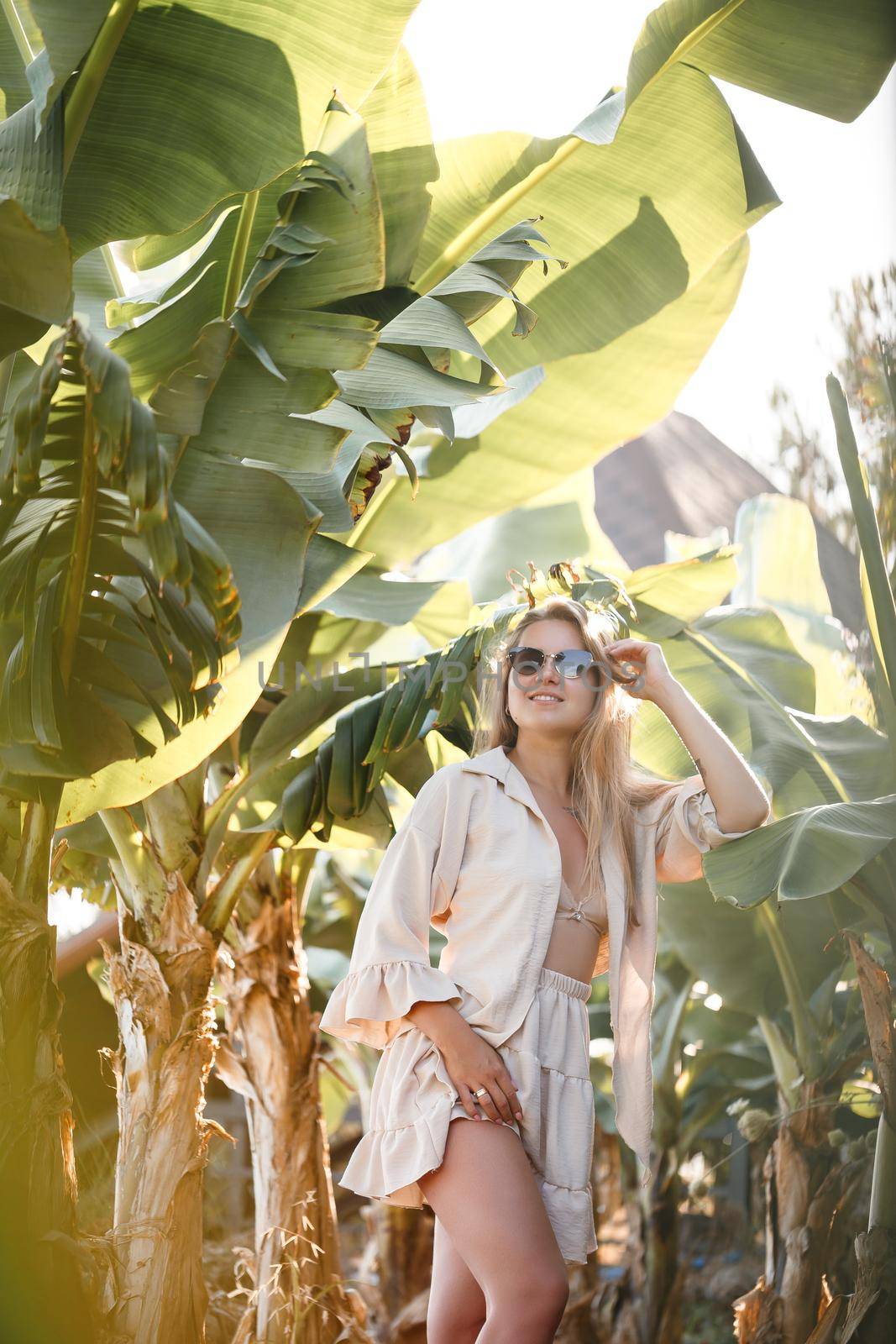 A beautiful young woman with long blond hair of European appearance stands near the banana trees. Girl in tropical forest on a sunny summer day. Selective focus by Dmitrytph