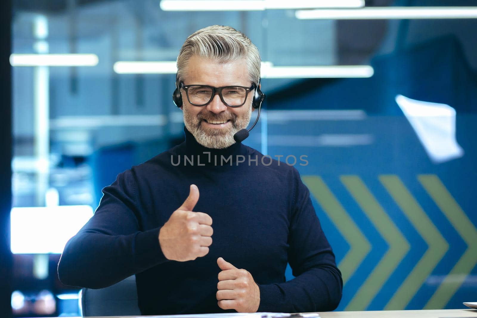 Video call senior gray haired man talking to colleagues remotely using headset smiling and looking at camera giving thumbs up, mature businessman inside modern office.