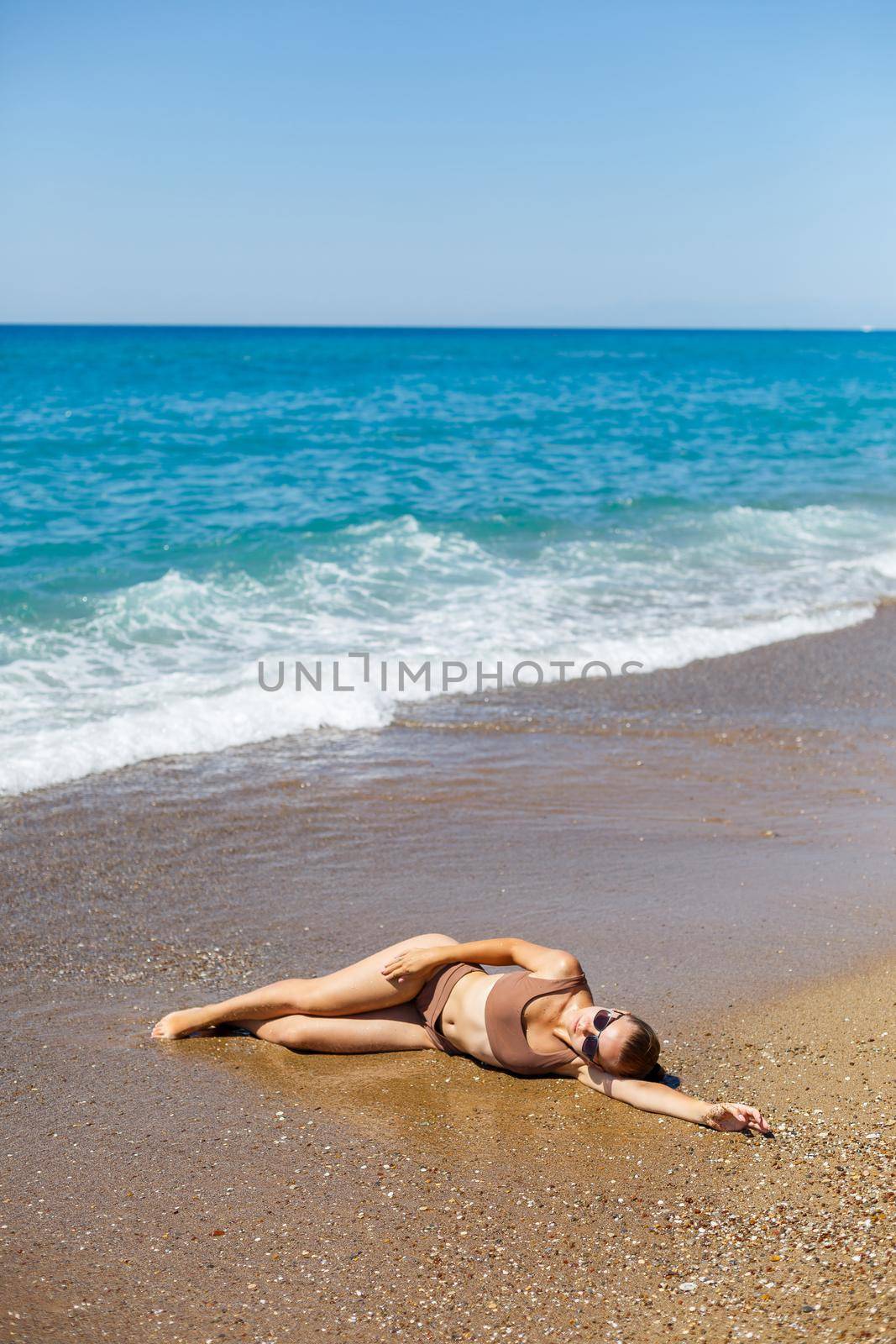 An attractive young woman in a bikini lies on the sand by the sea, relaxing on a deserted beach. Beautiful model in a swimsuit resting on the fine sand on a tropical island