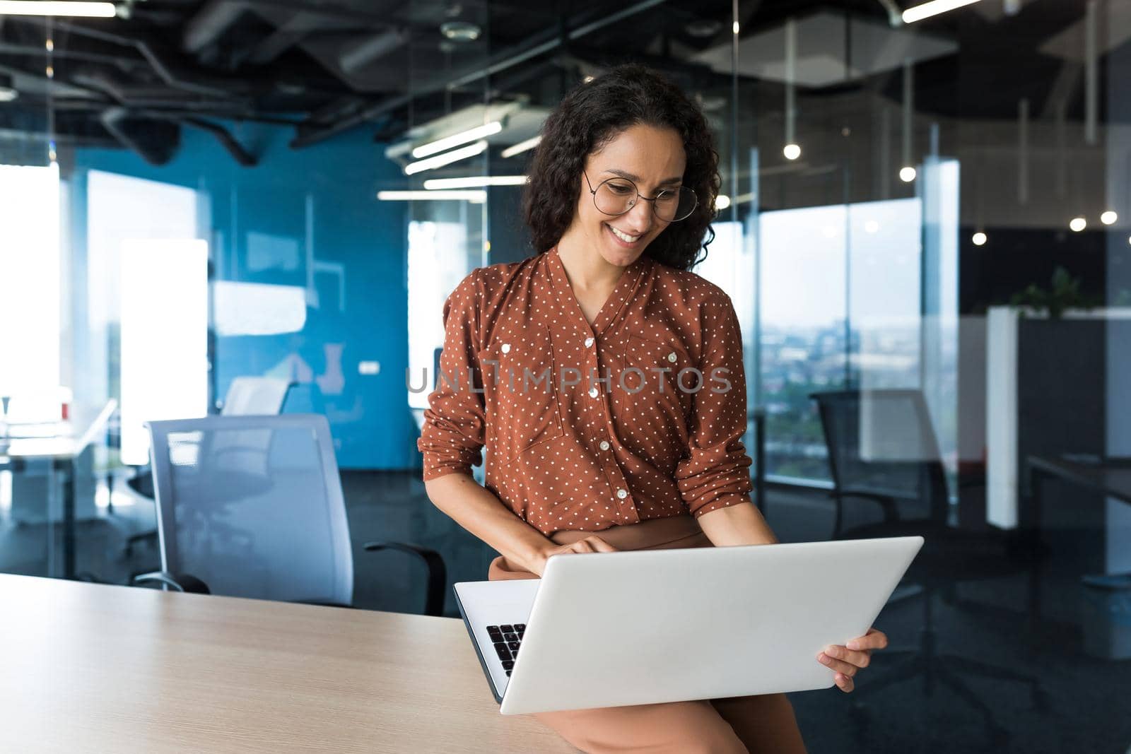 Young beautiful business woman working with laptop smiling and happy, Hispanic woman inside modern office building wearing glasses and curly hair.
