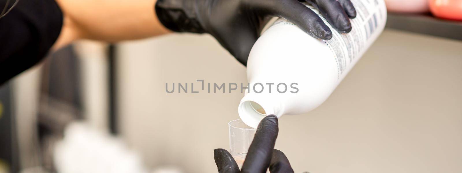 A hairdresser in black gloves is preparing hair dye with a bottle in a hair salon, close up