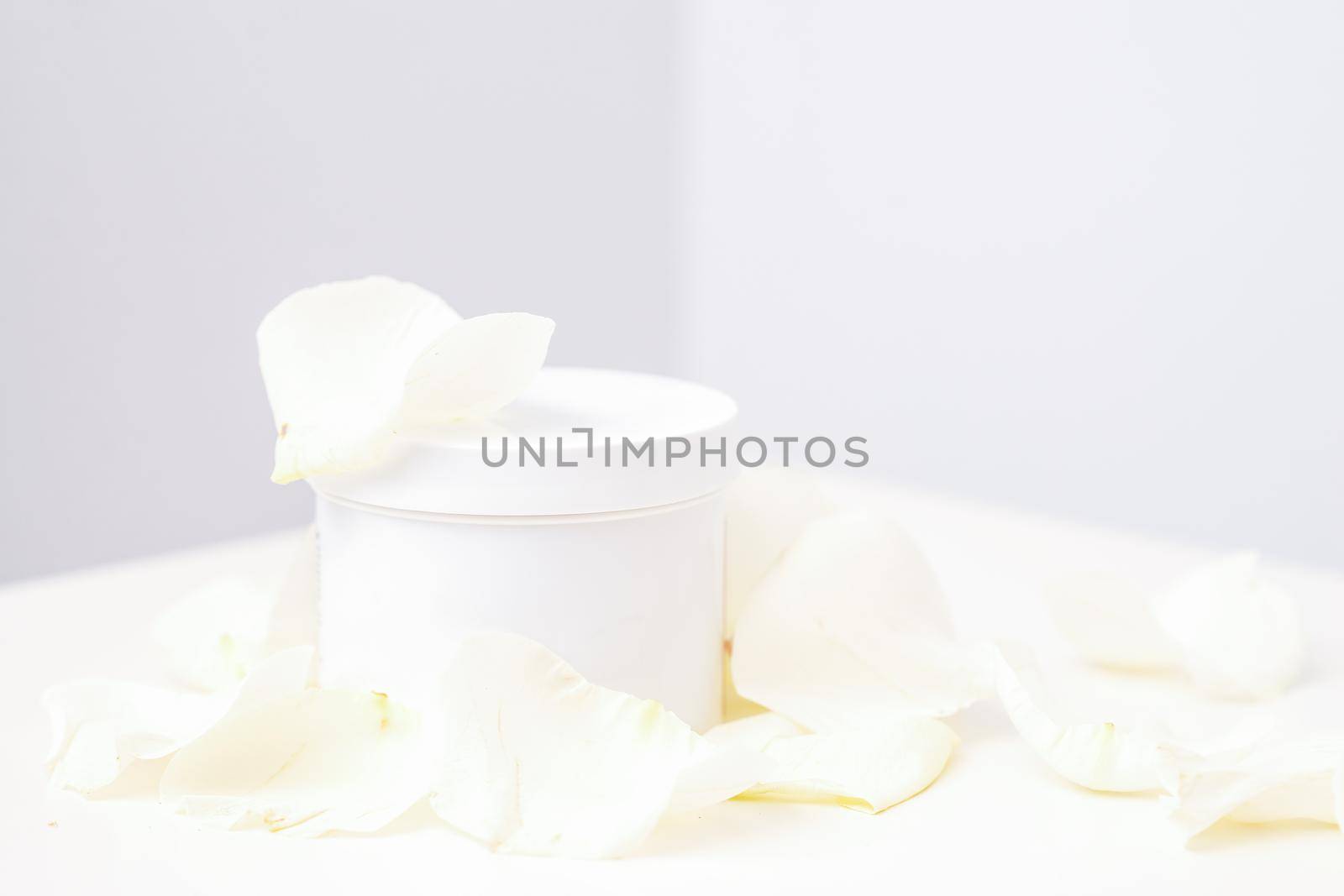 White jar with cream among delicate white rose flowers petals on a light background. Natural organic cosmetics concept, close up, mock-up