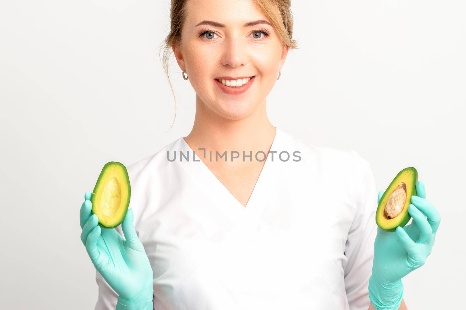 Portrait of smiling young female nutritionist doctor with organic avocado fruits posing at camera on white background, copy space. Benefits of proper nutrition