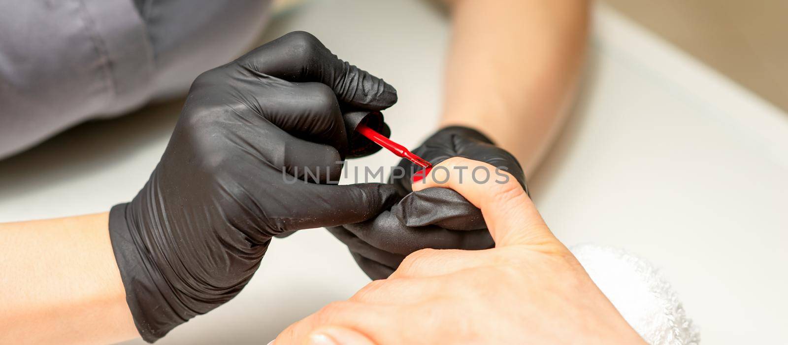 Manicure varnish painting. Close-up of a manicure master wearing rubber black gloves applying red varnish on a female fingernail in the beauty salon. by okskukuruza