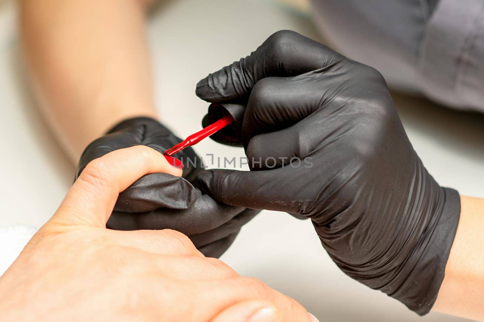 Manicure varnish painting. Close-up of a manicure master wearing rubber black gloves applying red varnish on a female fingernail in the beauty salon. by okskukuruza