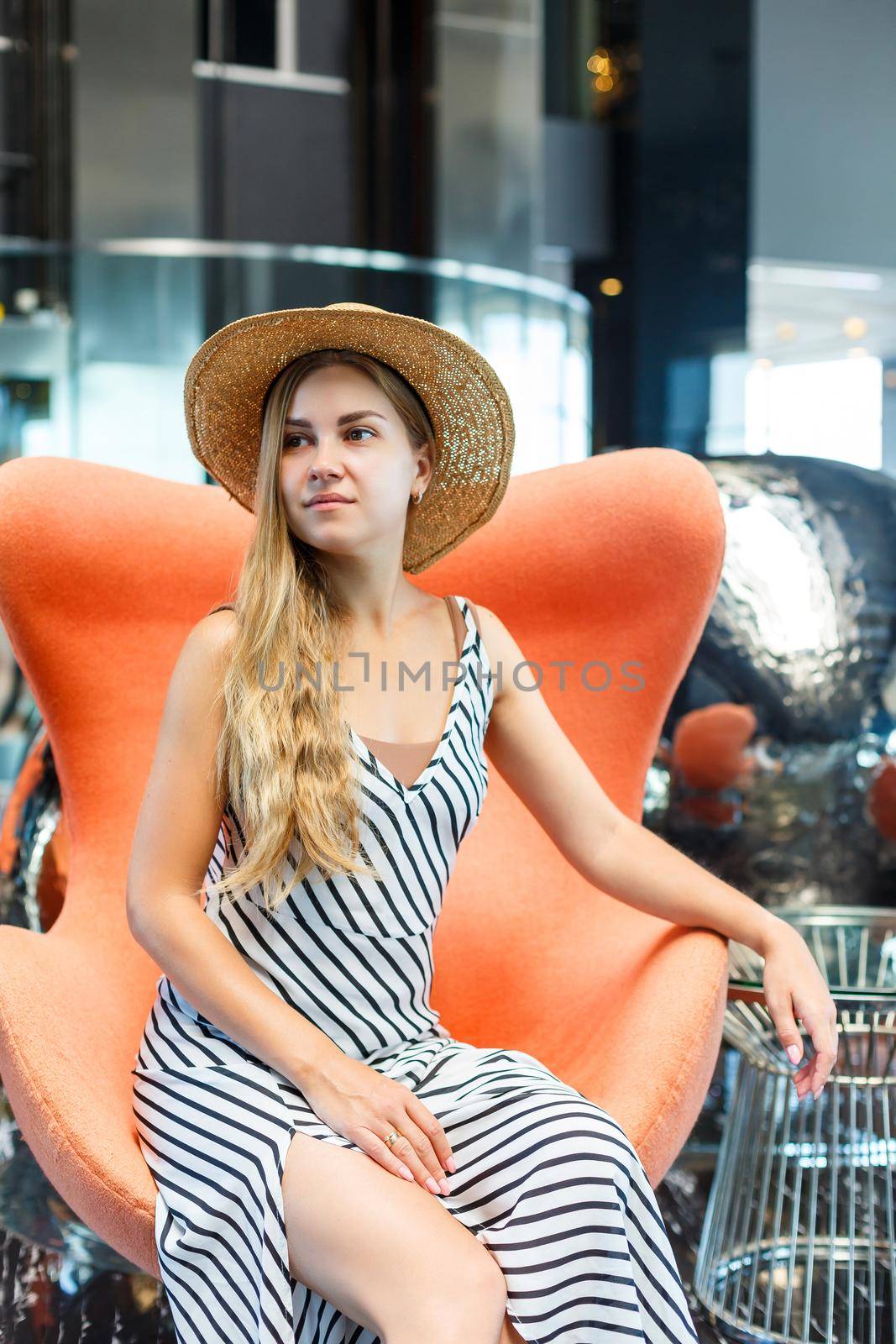 A beautiful young woman in a dress and a hat is sitting in the lobby of a luxury hotel. She is on vacation.