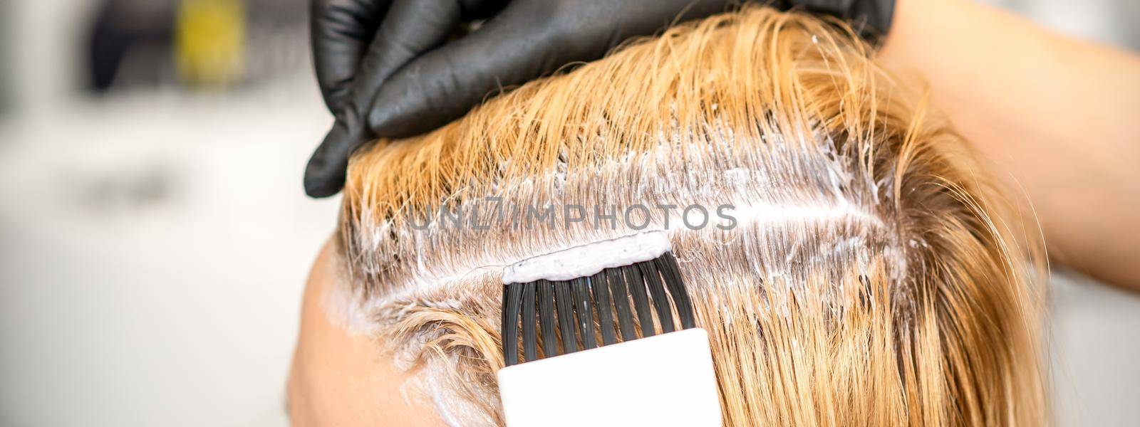 The hairdresser is dyeing blonde hair roots with a brush for a young woman in a hair salon. by okskukuruza