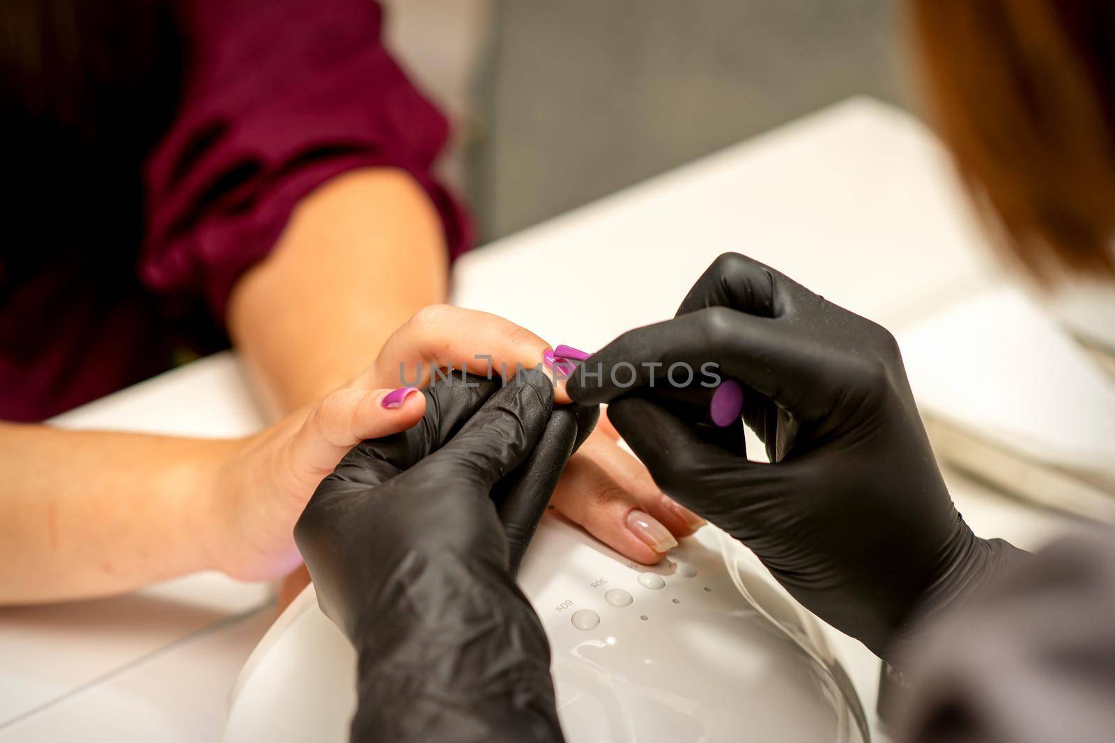 Professional manicure. A manicurist is painting the female nails of a client with purple nail polish in a beauty salon, close up. Beauty industry concept. by okskukuruza