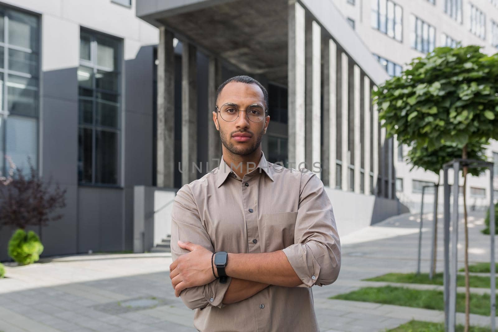 Serious and pensive young office worker with crossed arms looking at camera, businessman in shirt outside office building pensive.