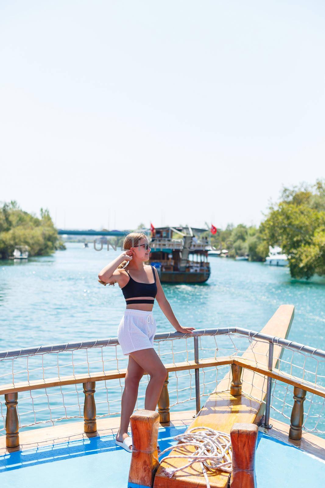 A young slender tanned woman in white shorts and a top, wearing sunglasses. Walk on a hot sunny day on a yacht along the river
