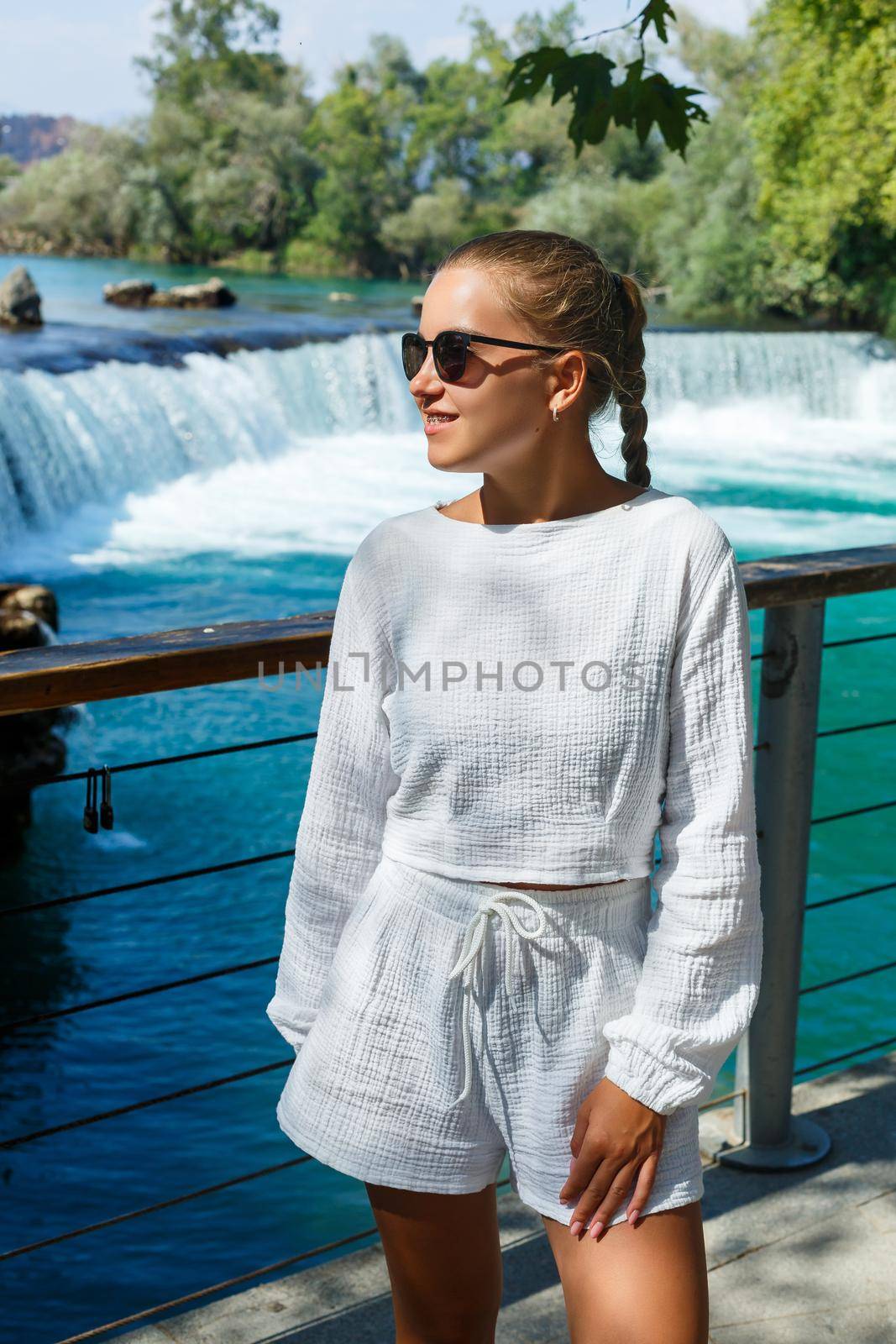 A beautiful young woman in white shorts and a shirt against the backdrop of a mountain waterfall against the backdrop of a dense forest. Beautiful landscape