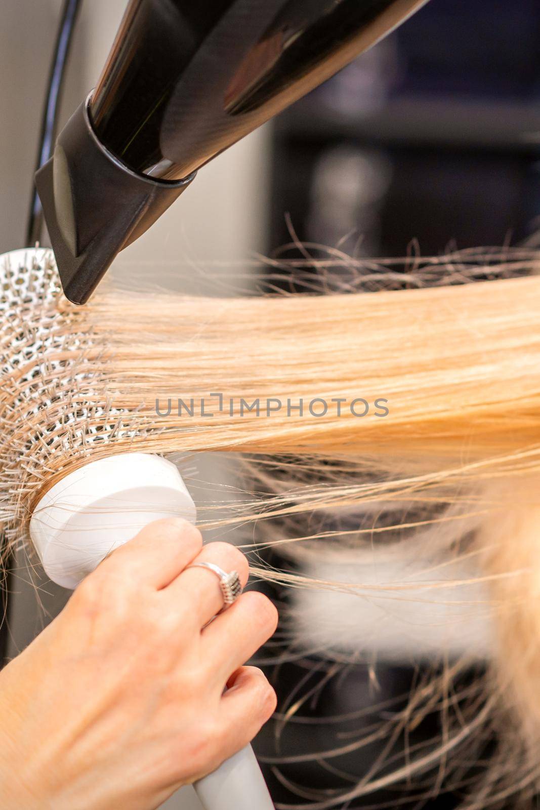Drying straight blond hair with black hairdryer and white round brush in hairdresser salon, close up. by okskukuruza