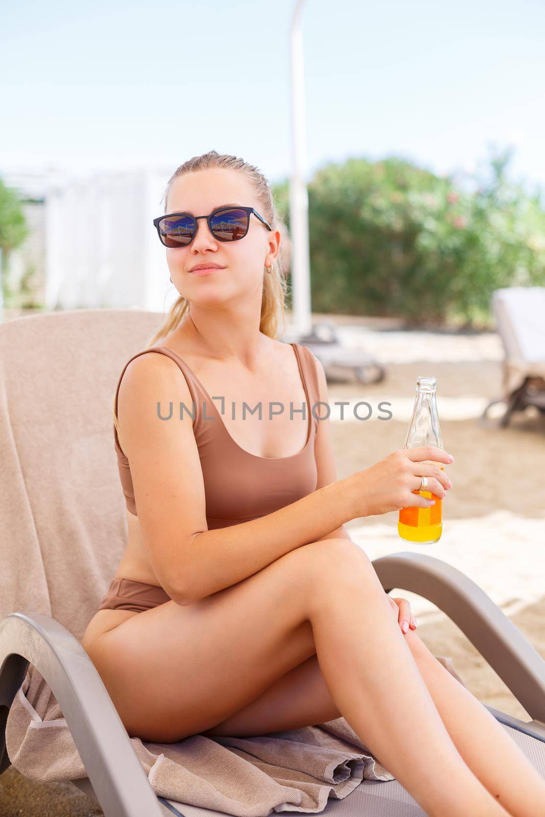 A beautiful slim woman in a beige swimsuit sits on a sun lounger with a bottle of refreshing water and enjoys her vacation by the sea