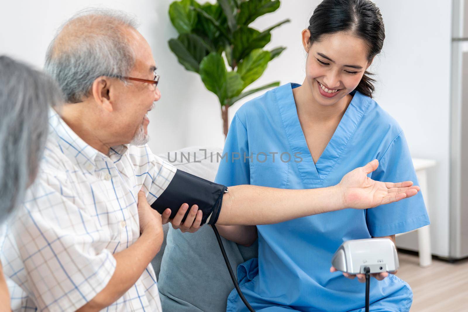An elderly man having a blood pressure check by his personal caregiver with his wife sitting next to him in their home.