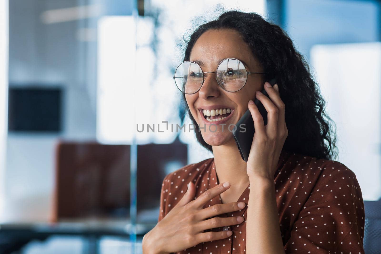 Young and beautiful hispanic woman talking on the phone and smiling happy, business woman close up in glasses and curly hair working inside a modern office building