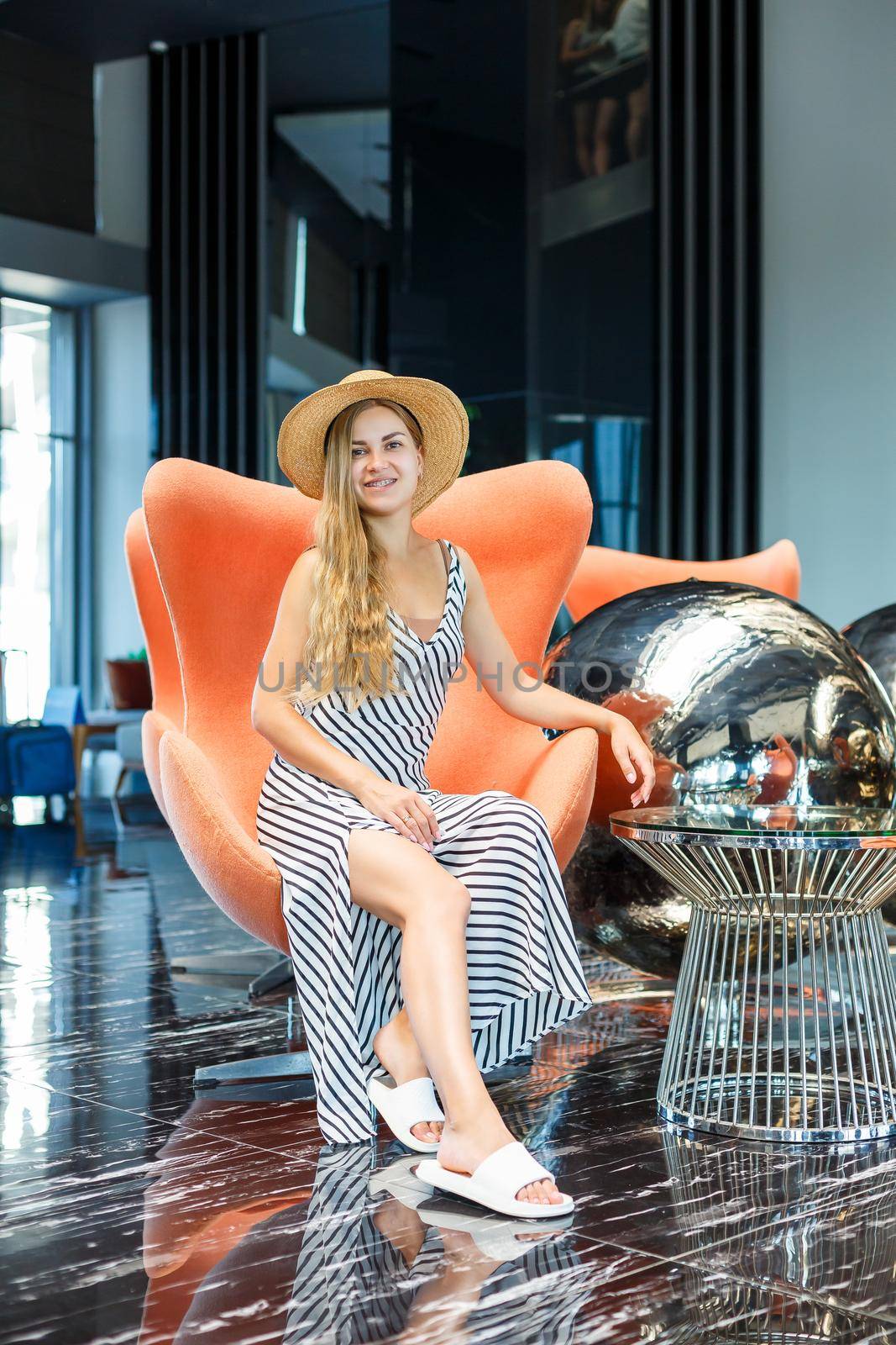 A beautiful young woman in a dress and a hat is sitting in the lobby of a luxury hotel. She is on vacation.