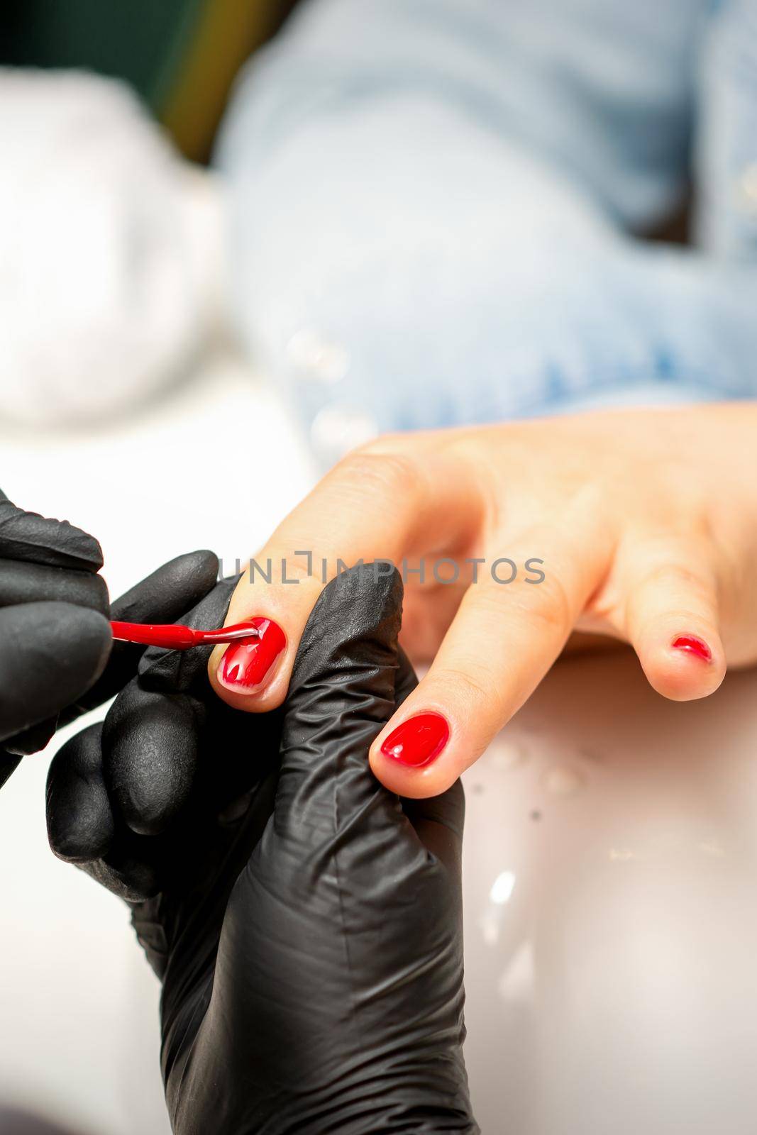 Manicure varnish painting. Close-up of a manicure master wearing rubber black gloves applying red varnish on a female fingernail in the beauty salon. by okskukuruza