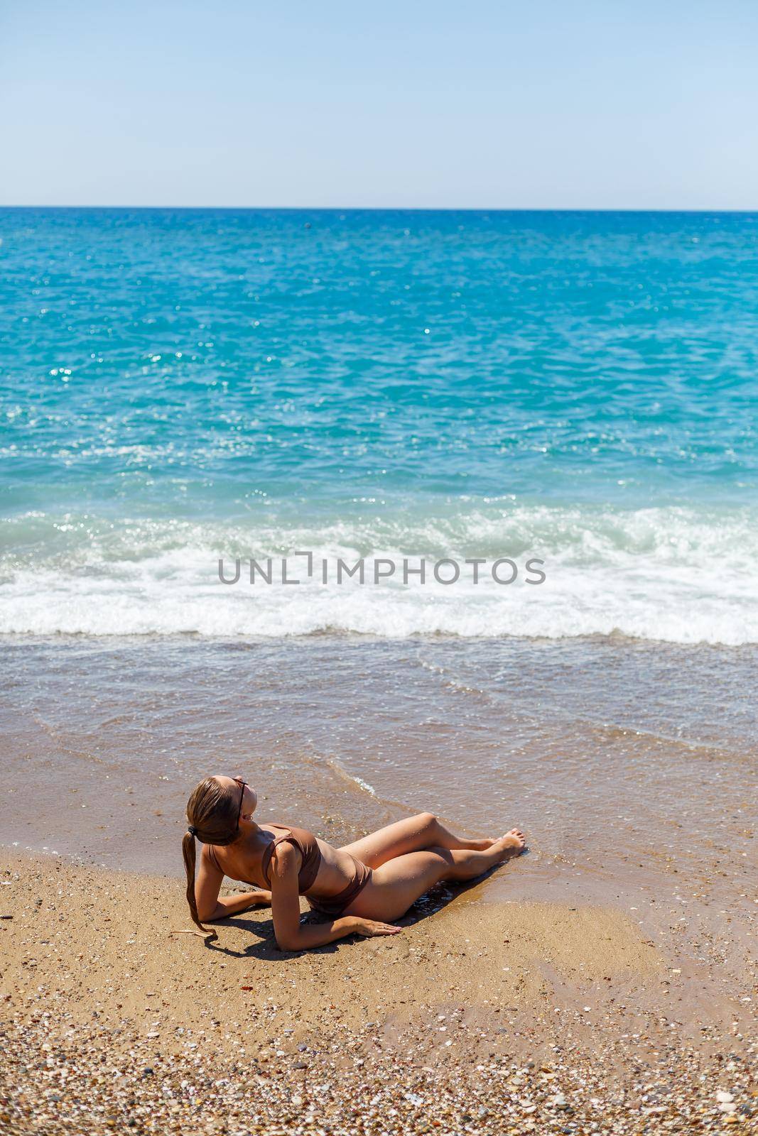 An attractive young woman in a bikini lies on the sand by the sea, relaxing on a deserted beach. Beautiful model in a swimsuit resting on the fine sand on a tropical island by Dmitrytph