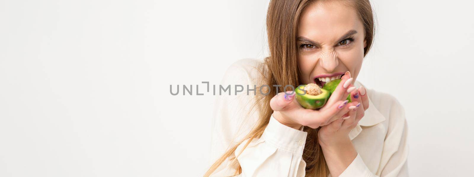 Portrait of a lovely smiling young brunette caucasian woman wearing the white shirt with long hair holding and showing avocado, standing isolated over white background