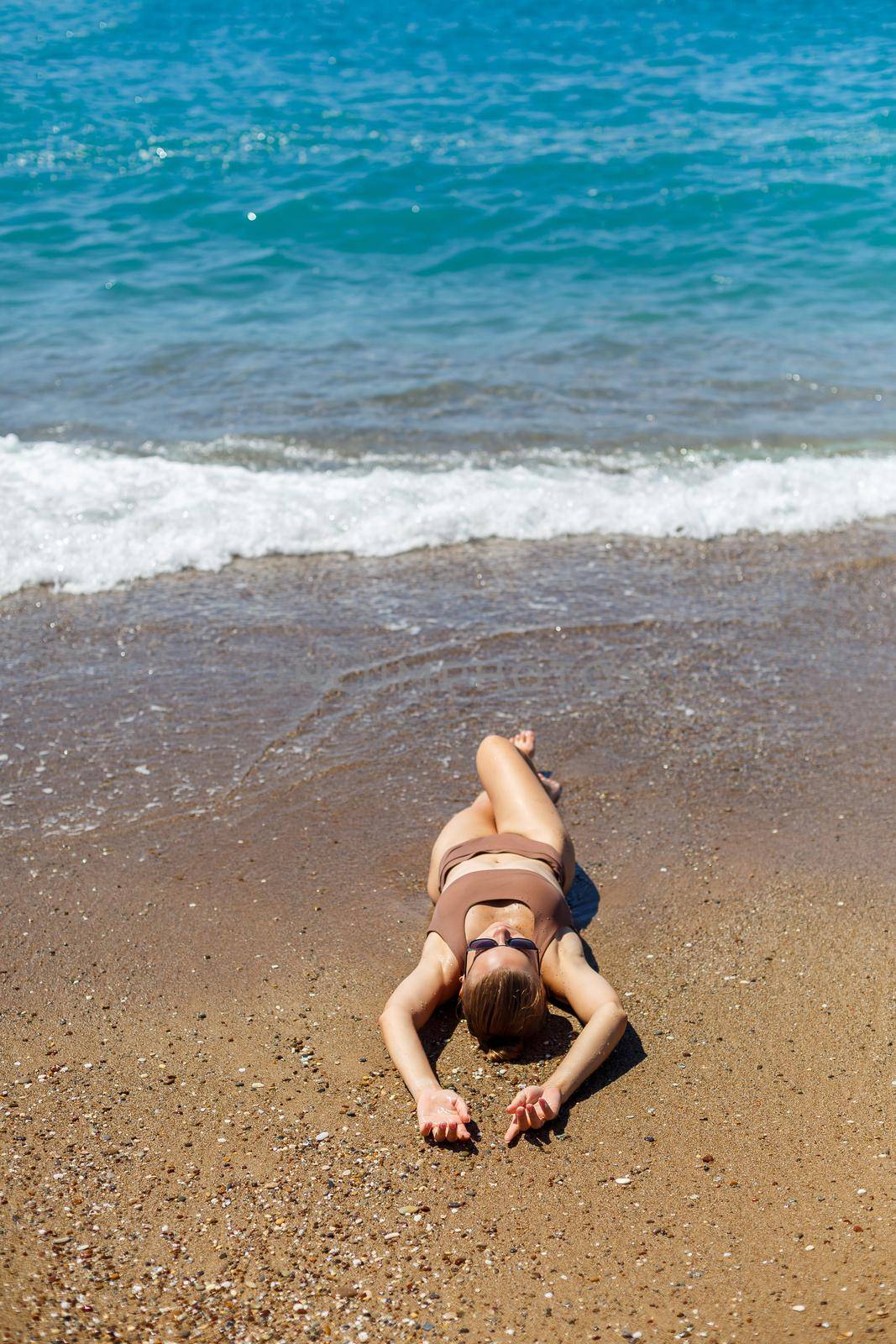 An attractive young woman in a bikini lies on the sand by the sea, relaxing on a deserted beach. Beautiful model in a swimsuit resting on the fine sand on a tropical island by Dmitrytph