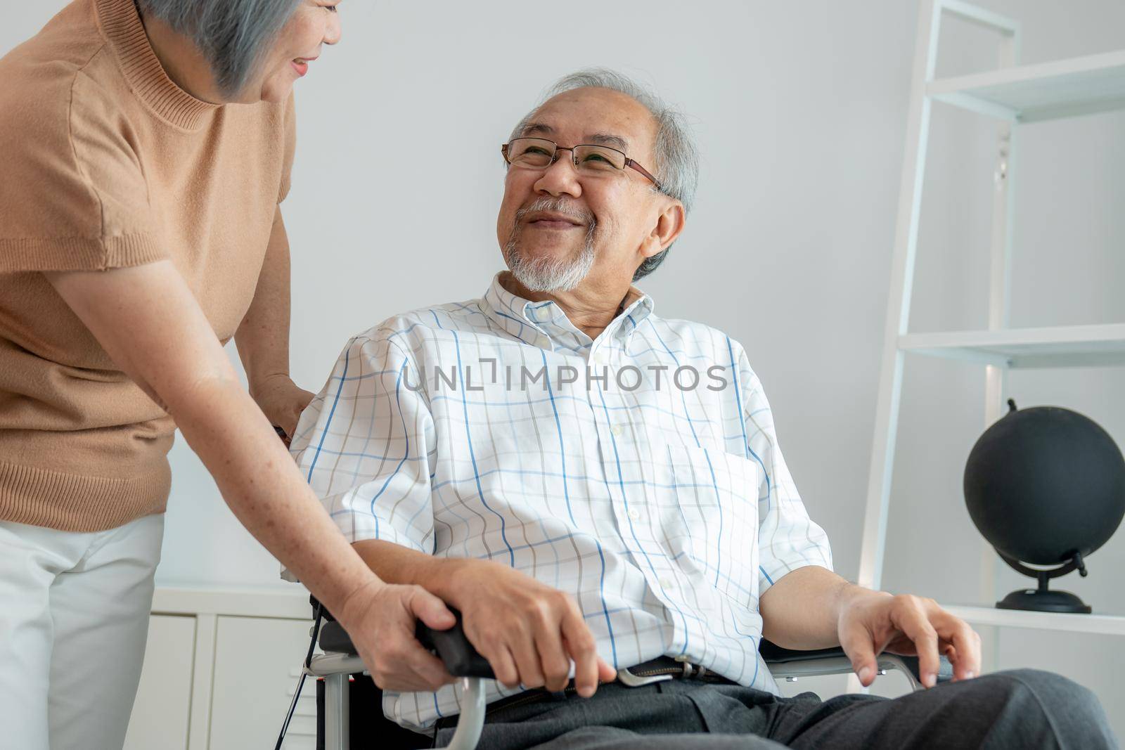 Senior wife giving support to her husband in his wheelchair with love, contented pensioner life. A senior couple is understanding and smiling at each other.