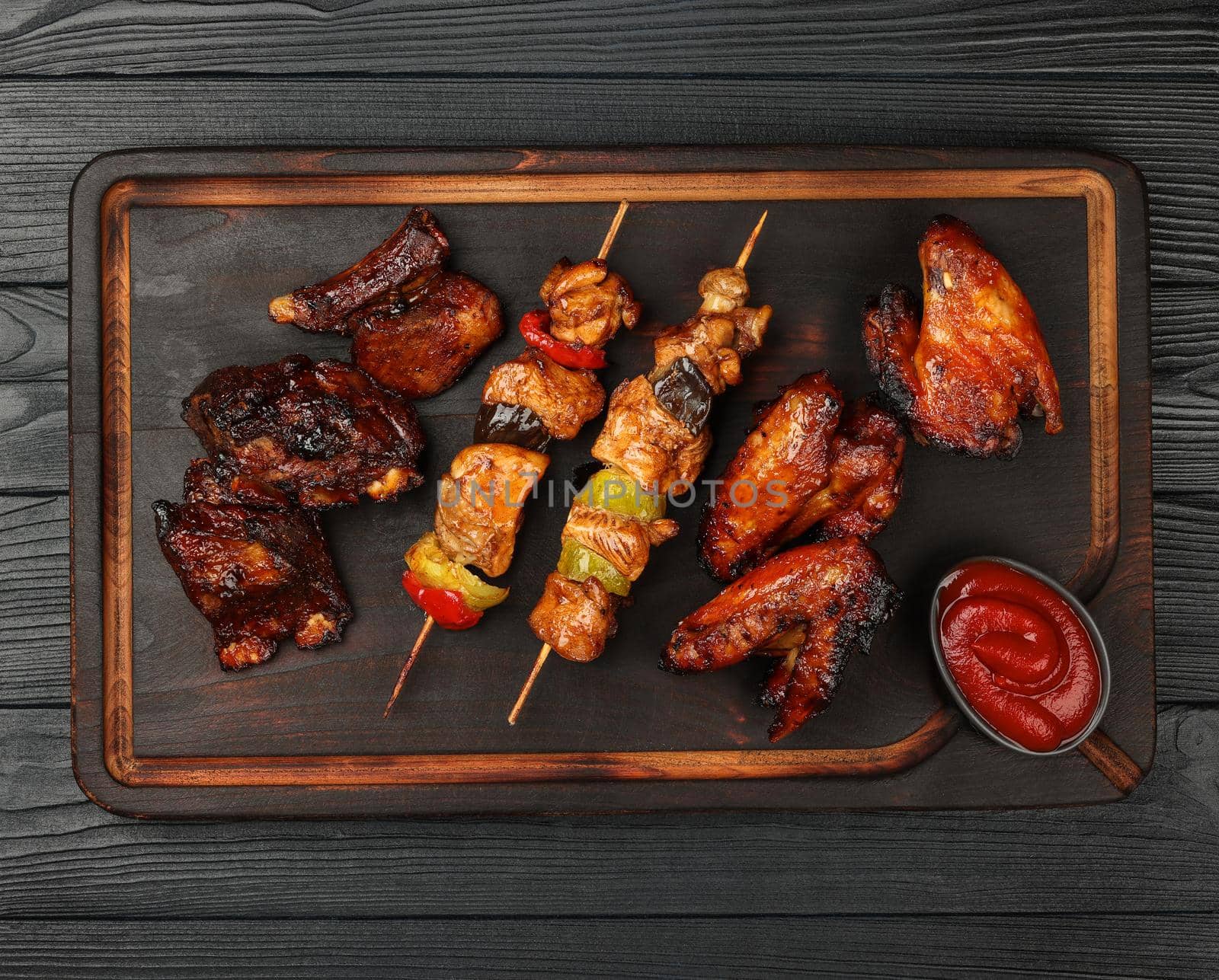 Close up portion of mixed grilled barbecue combo plate with chicken buffalo wings, beef spare ribs and skewer meat on wooden board over black table planks, elevated top view, directly above