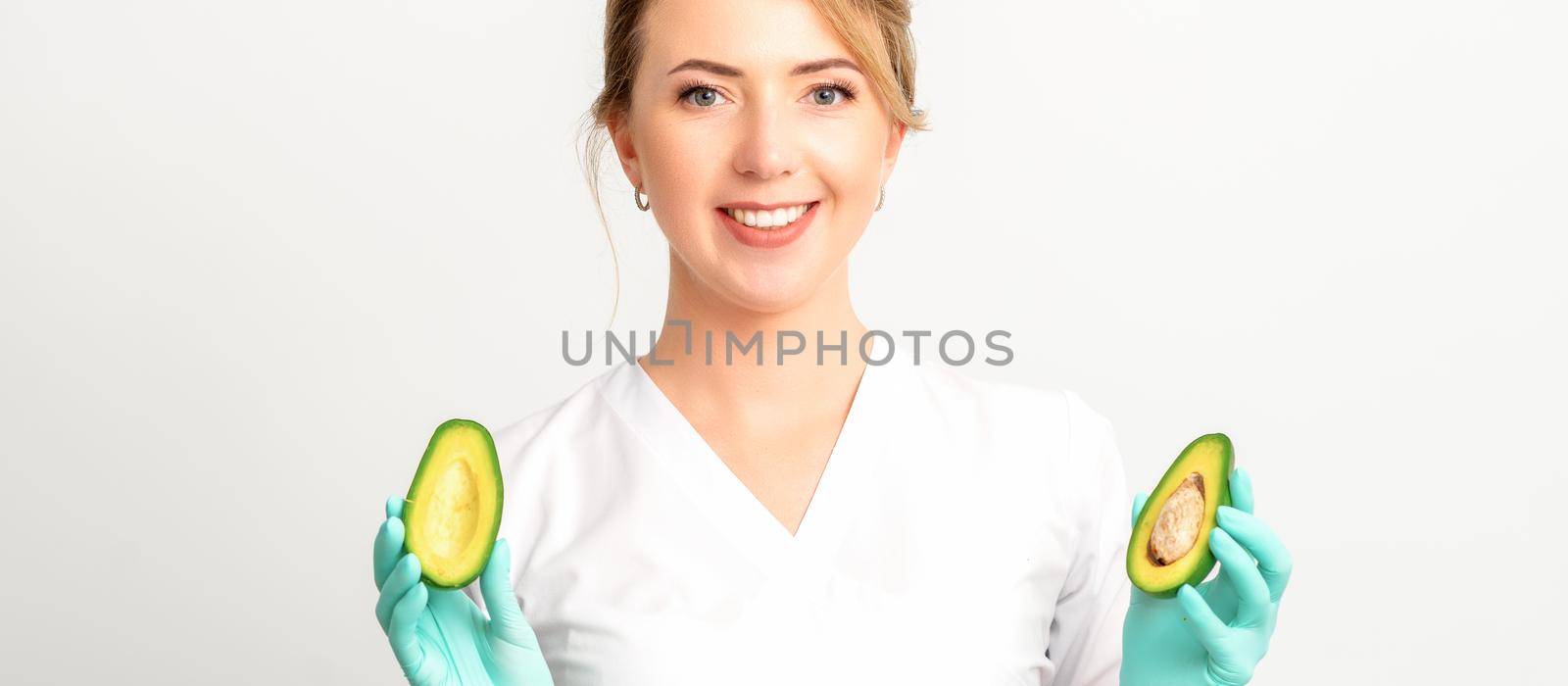 Portrait of smiling young female nutritionist doctor with organic avocado fruits posing at camera on white background, copy space. Benefits of proper nutrition. by okskukuruza