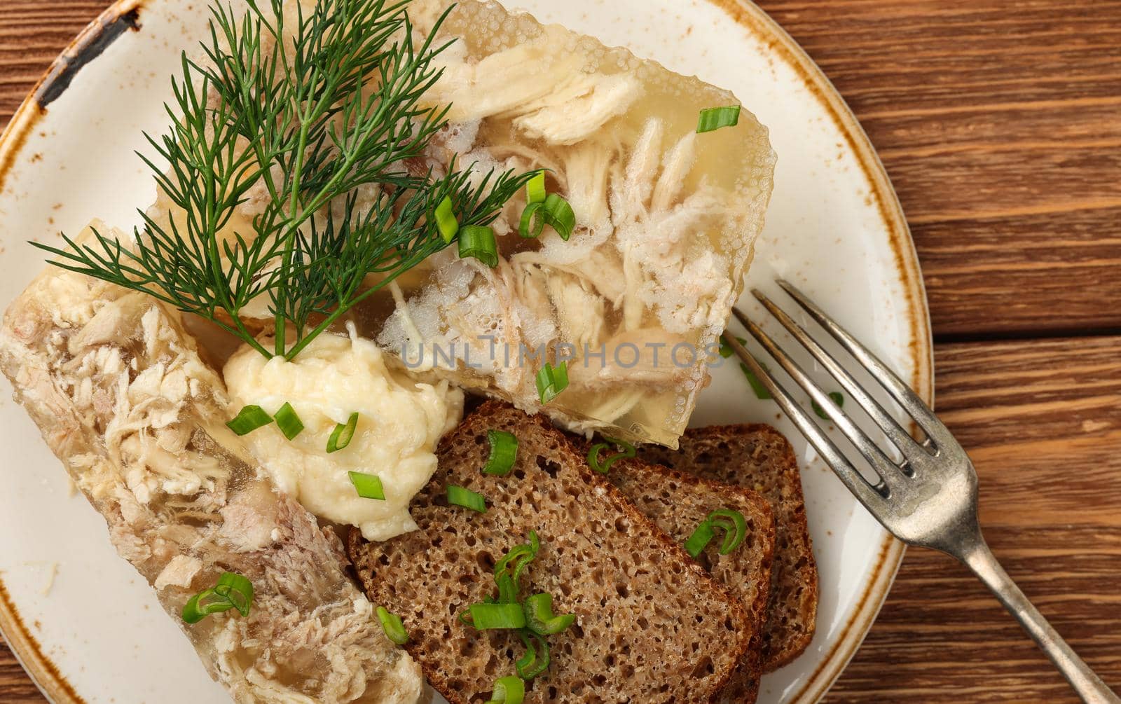 Close up portion of aspic or traditional Eastern European chicken meat jelly with slices of rye bread and horseradish dressing sauce, on brown wooden table, elevated top view, directly above