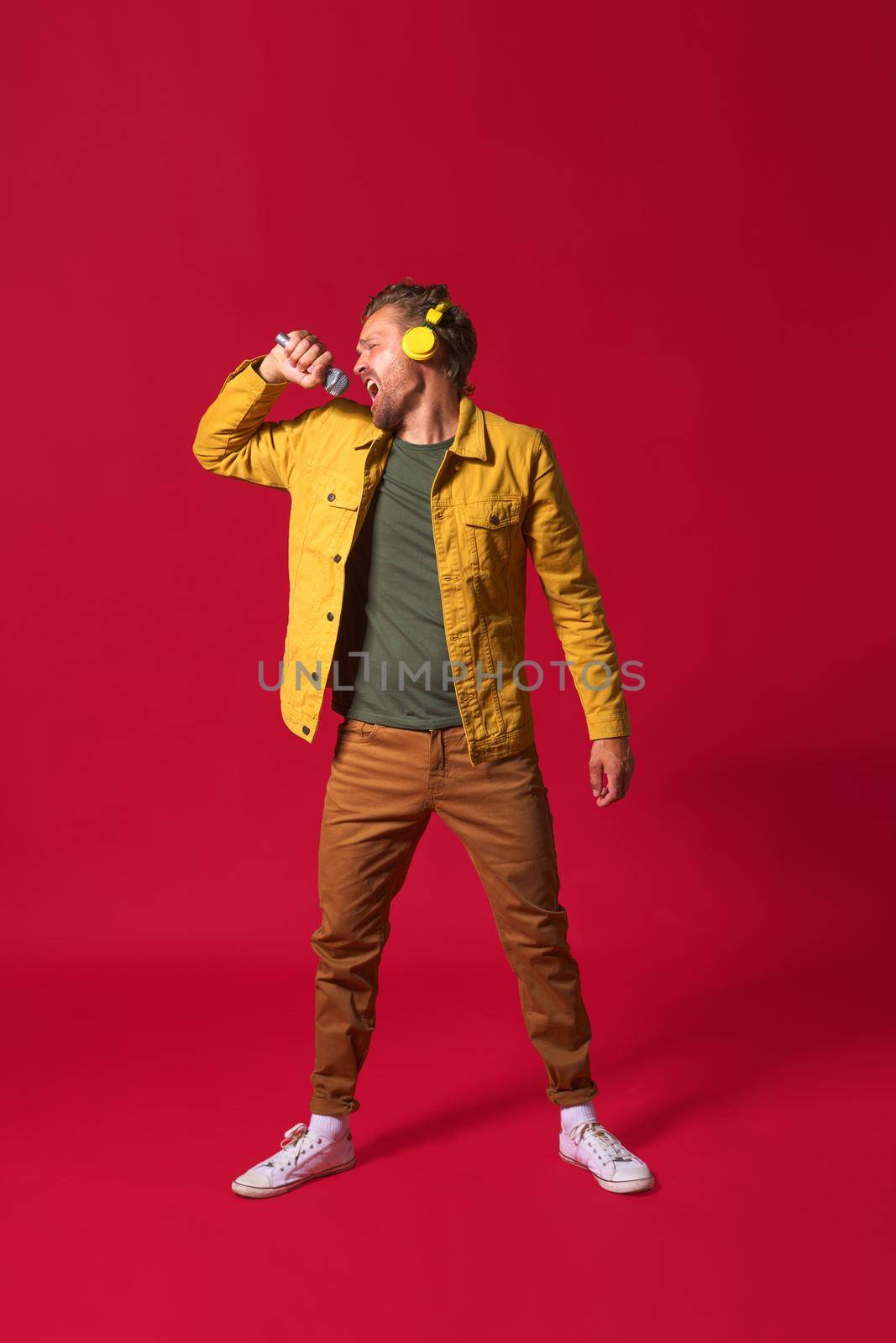 Singing young man enjoying his favorite song holding microphone and wireless headphones wearing casual jacket isolated on red background. Man sing while recording his voice. A star is born.