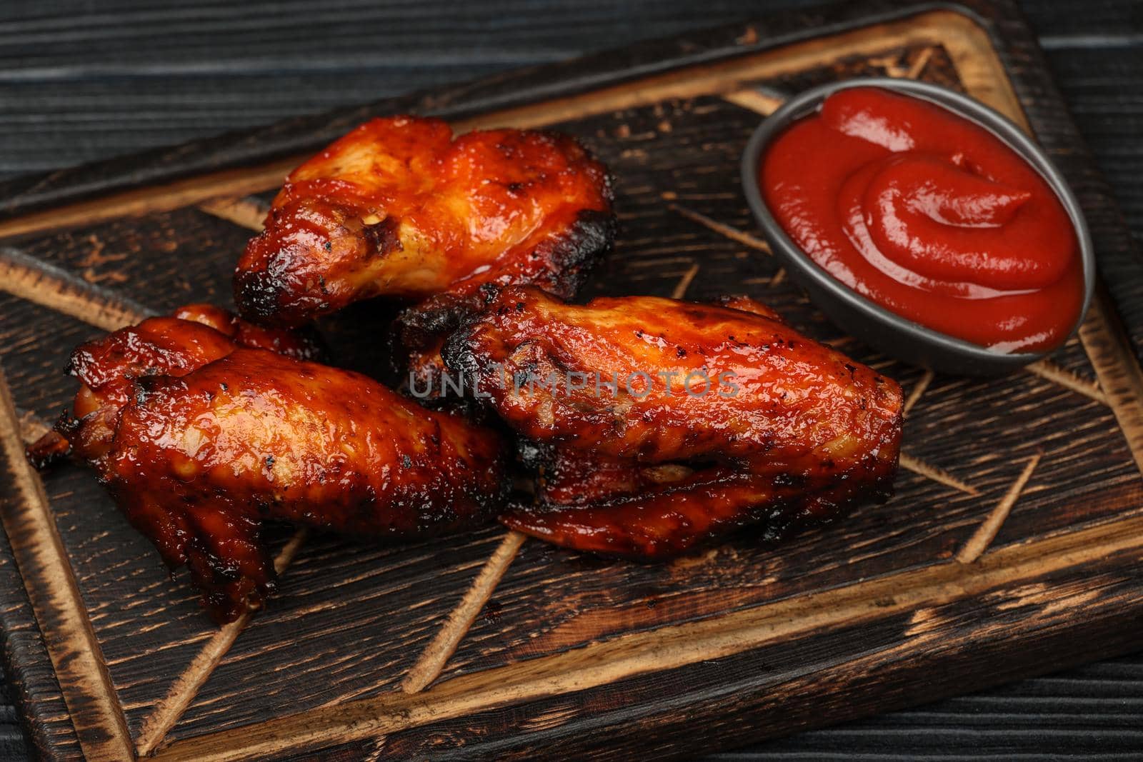 Close up portion of grilled barbecue chicken buffalo wings and BBQ tomato sauce served on wooden board over black table planks, high angle view, personal perspective