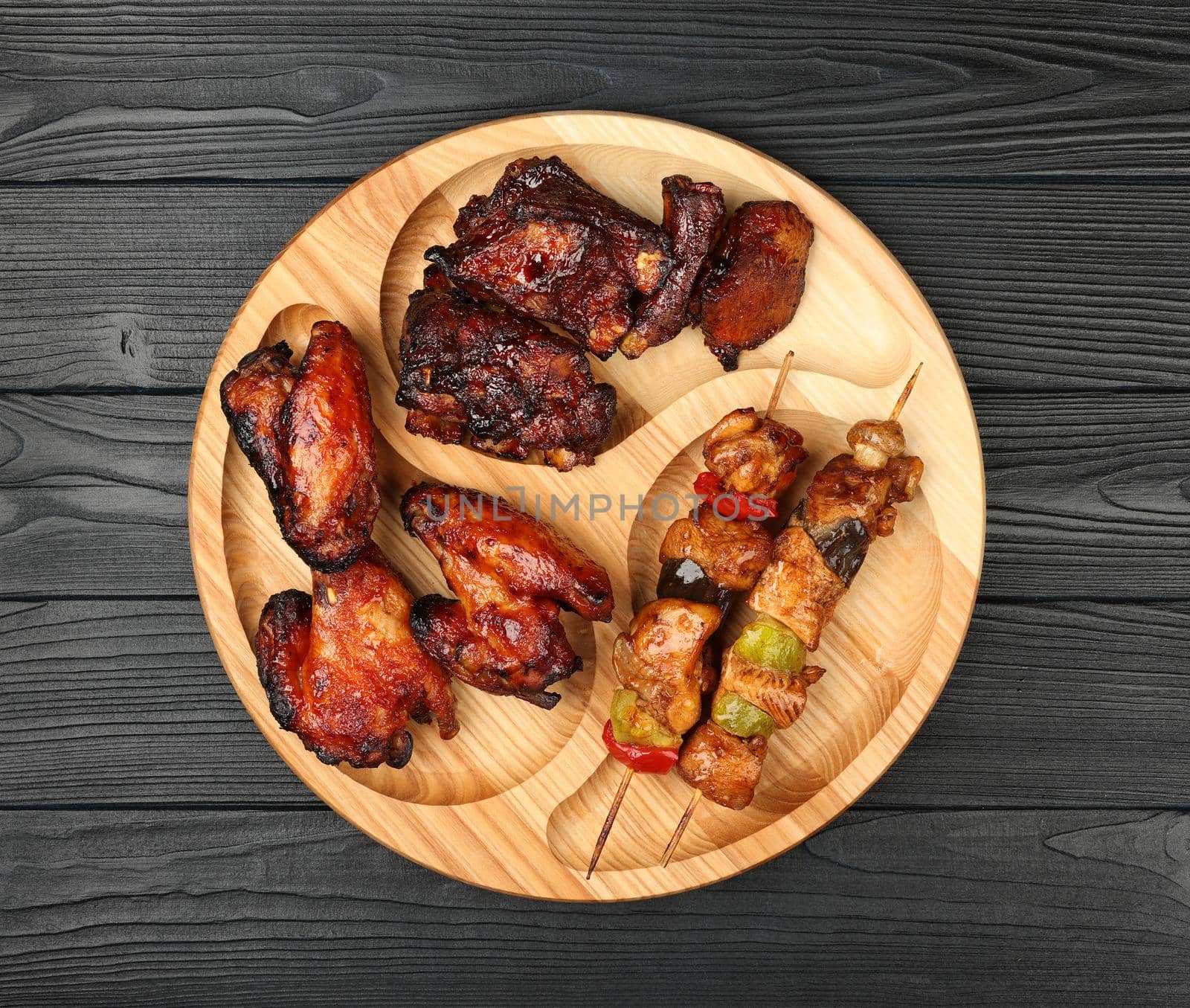 Close up portion of mixed grilled barbecue combo plate with chicken buffalo wings, beef spare ribs and skewer meat on wooden board over black table planks, elevated top view, directly above