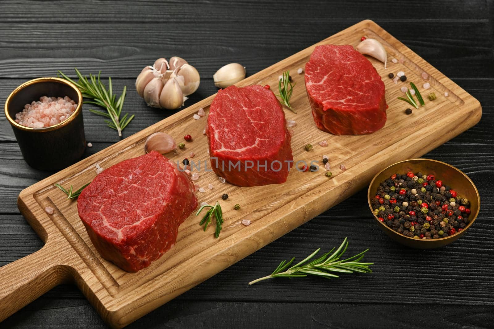Close up three aged prime marbled raw tenderloin or fillet mignon beef steaks on brown oak wood cutting board, with spices, over black wooden table background, high angle view