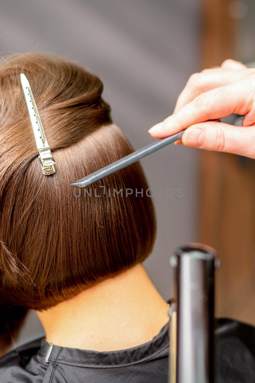 Hairstylist is straightening short hair of young brunette woman with a flat iron in a hairdresser salon, close up. by okskukuruza