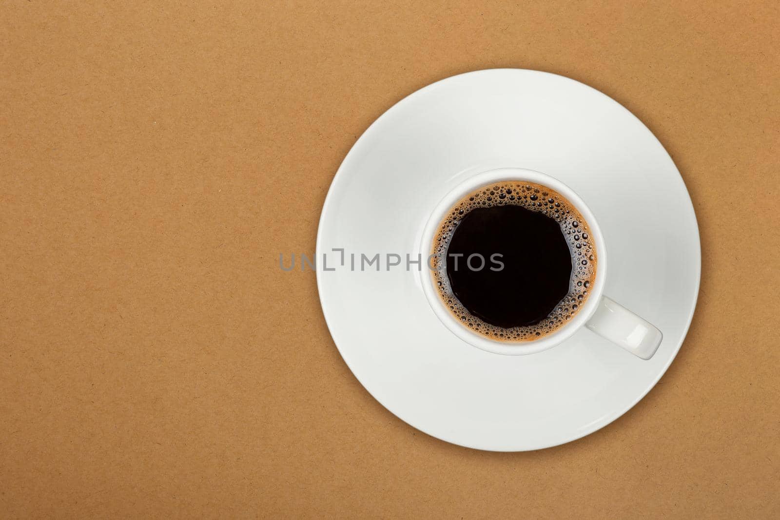 Close up one white cup full of black coffee on saucer over brown paper parchment background, elevated top view, directly above